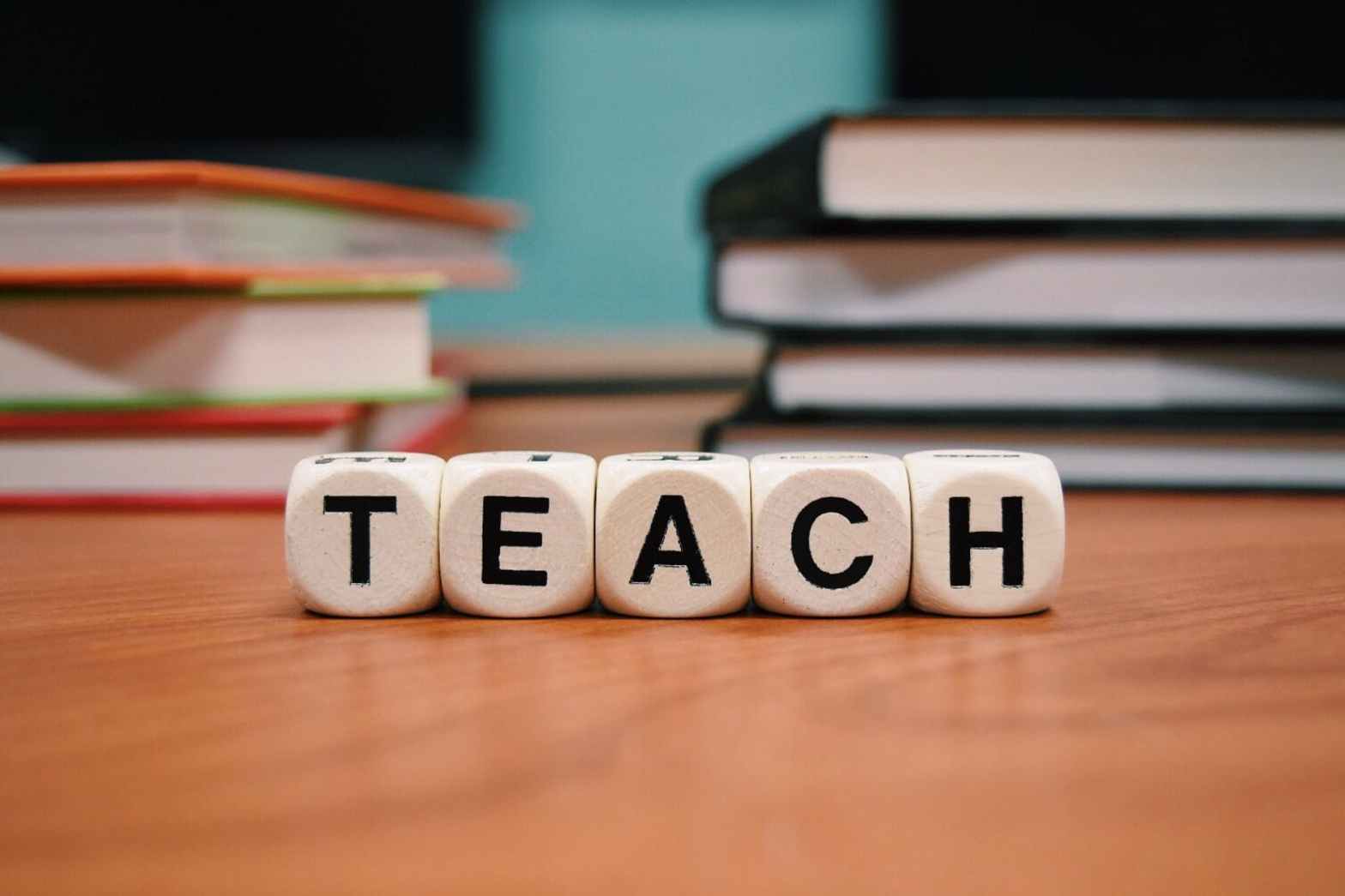 Wooden lettered blocks spelling ‘TEACH’ on a classroom desk, with stacks of books in the background—symbolizing education, instruction, and fostering critical thinking in children.