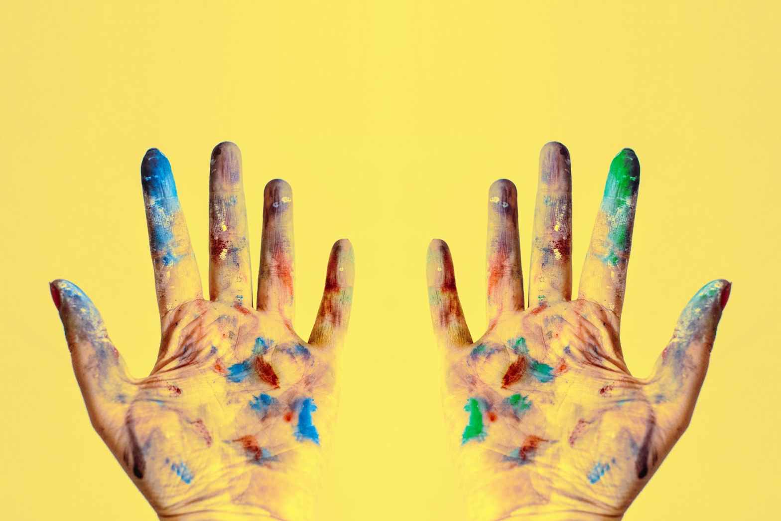 Two open hands covered in colorful paint against a bright yellow background, symbolizing creativity, self-expression, and the artistry of leadership.